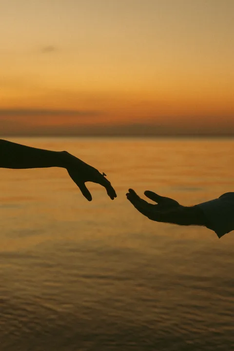 Two hands reaching toward each other over the water at sunset